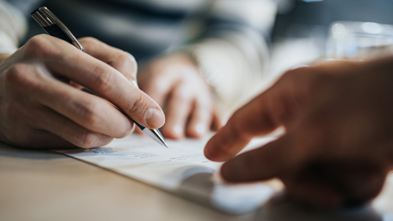Close up of unrecognizable man signing a contract while financial advisor is aiming at the place he need to sign.