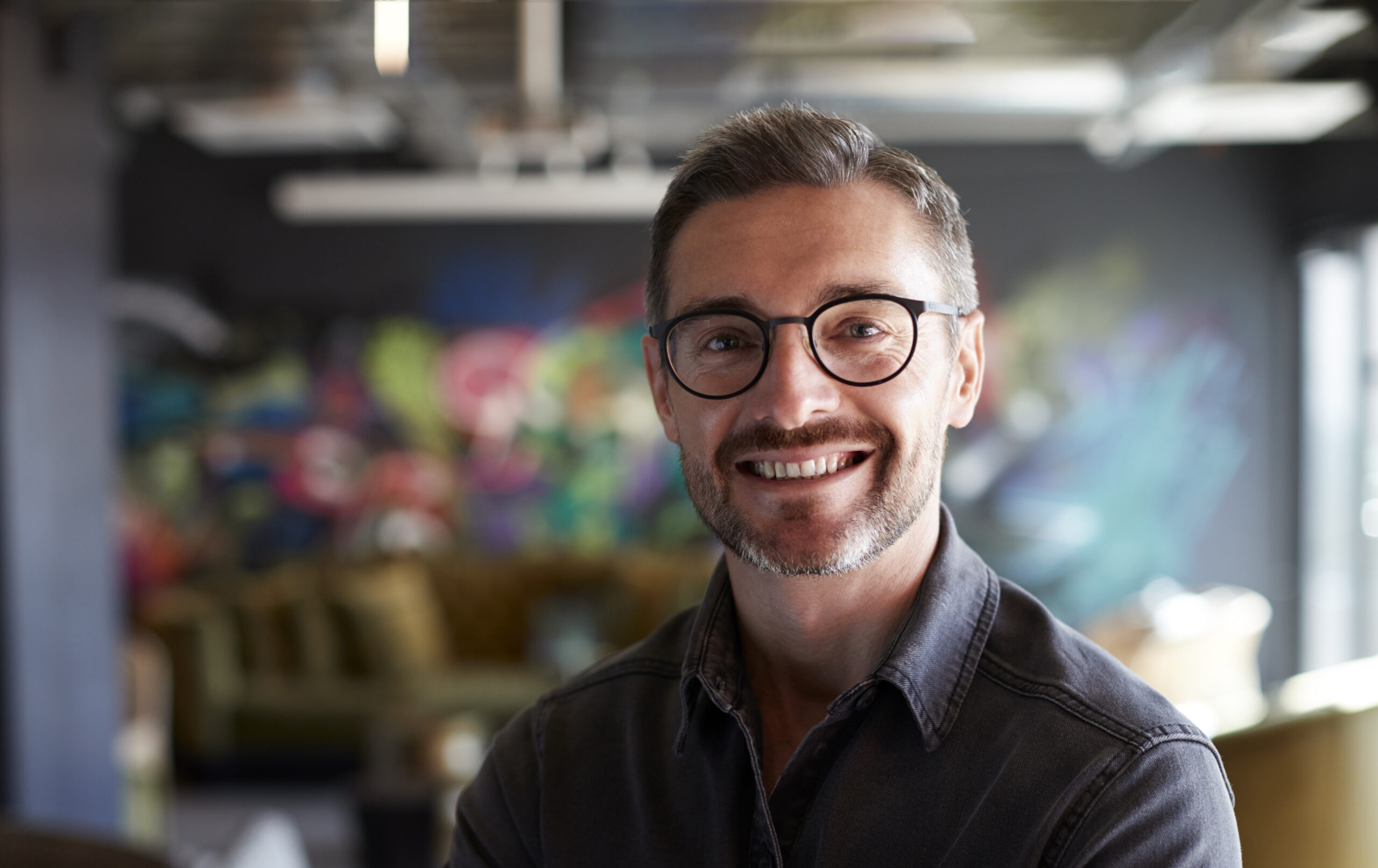 Middle aged white male creative in casual office lounge area looks to camera smiling, close up